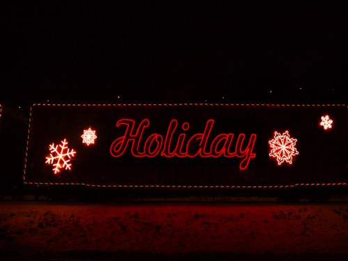 A brightly lit train car displays the word "Holiday" with snowflake decorations against a dark background.