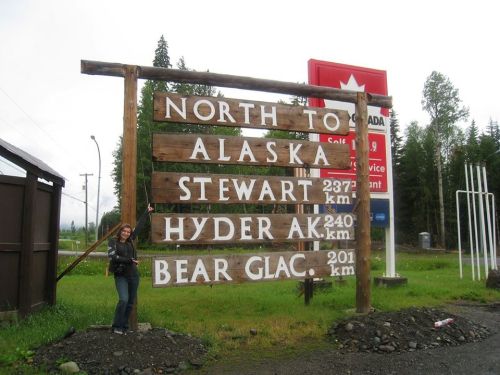 A wooden signpost indicating distances to Alaska, Stewart, Hyder, and Bear Glacier, with a person standing beside it.