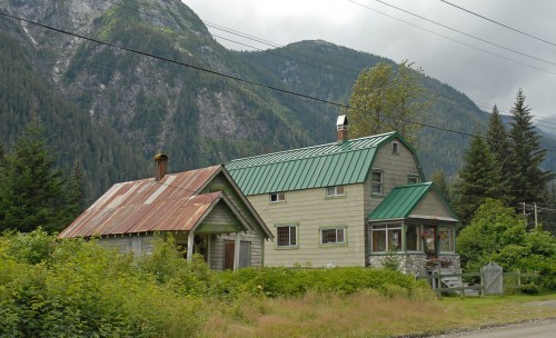 A rustic house with a green metal roof, surrounded by greenery and mountains under a cloudy sky.