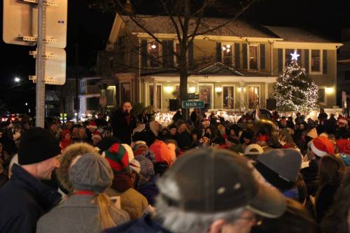 A crowd gathers at night for a festive event, with a decorated house and Christmas tree in the background.
