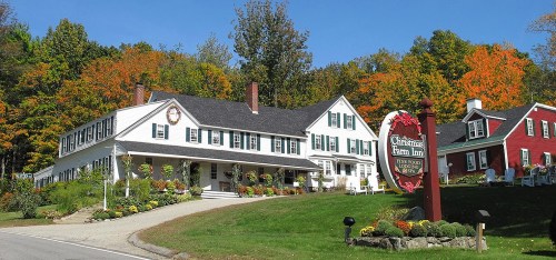 A charming inn with white siding and green shutters, surrounded by colorful autumn trees and a welcoming sign.