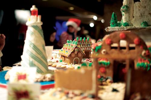 A festive display of decorated gingerbread houses and holiday treats on a table, with a person in a Santa hat in the background.