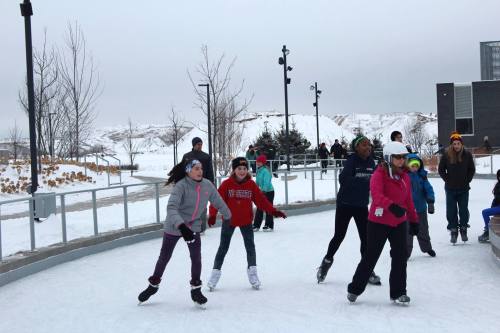 A group of children and adults ice skating on an outdoor rink surrounded by snow and trees on a cloudy day.