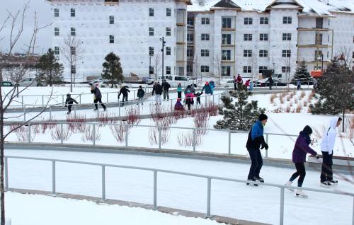 People ice skating on a winding rink surrounded by snow and a partially constructed building in the background.