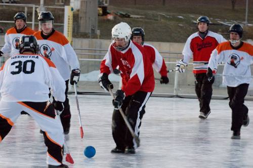 A group of players in hockey gear competes on an ice rink, with one player dribbling a blue ball.