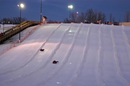 Two sledders descend a snowy hill at dusk, with lights illuminating the slope and a wooden structure in the background.