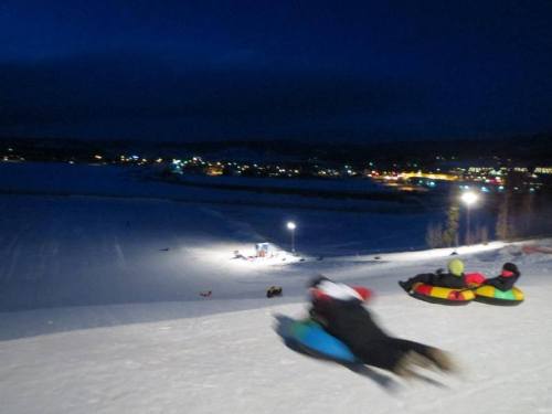 A snowy hill at night with people tubing down, colorful tubes, and lights illuminating the scene in the background.