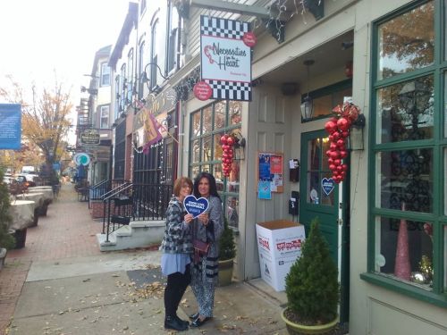 Two women stand outside a charming storefront decorated with holiday ornaments, holding heart-shaped signs.