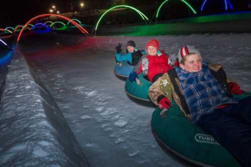 Three children in winter gear enjoy tubing down a snowy slope, illuminated by colorful lights in the background.