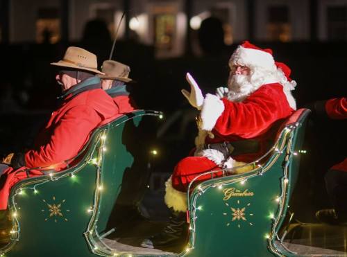 Santa Claus waves from a decorated sleigh, accompanied by another person in a hat, during a festive event.