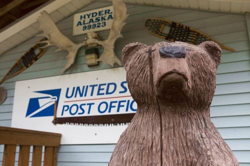 A wooden bear sculpture in front of the Hyder, Alaska Post Office, with decorative moose antlers above.