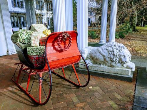 A red sleigh with wrapped gifts sits beside a stone lion statue on a brick path, surrounded by columns and trees.