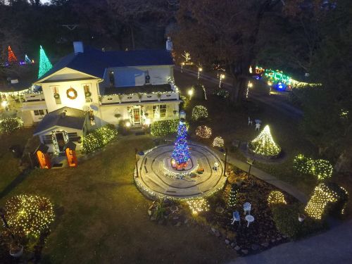 Aerial view of a beautifully decorated house with colorful Christmas lights and a central tree in a festive garden.