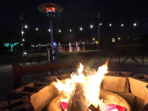 A cozy fire pit with flames, surrounded by outdoor seating and festive lights in the background at night.