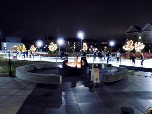 A nighttime ice skating rink with people skating and trees lit with holiday lights, featuring a fire pit in the foreground.