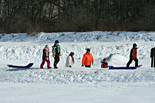 A group of people walking on a snowy path, some carrying sleds, with trees in the background.