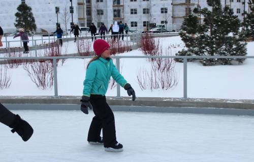 A girl in a teal jacket and pink hat ice skates on a rink surrounded by snow and trees.
