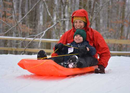 A smiling child and adult sled down a snowy hill, surrounded by trees, enjoying a winter day.