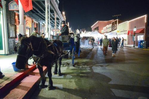 A horse-drawn carriage waits on a festive street at night, with people walking and holiday lights in the background.