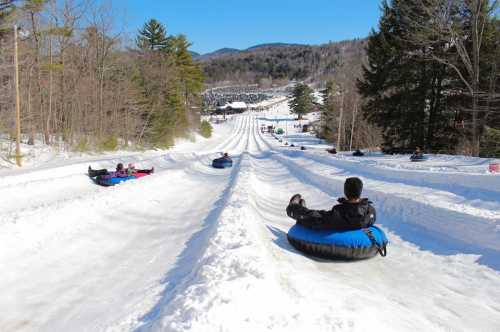 People tubing down snowy slopes on a sunny day, surrounded by trees and distant hills.