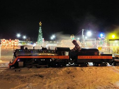 A steam train decorated with lights, with a person on board, surrounded by festive holiday decorations at night.