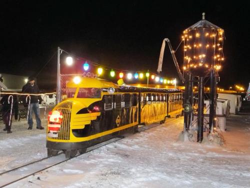 A brightly lit yellow train at night, decorated with colorful lights, near a vintage water tower. Snow covers the ground.
