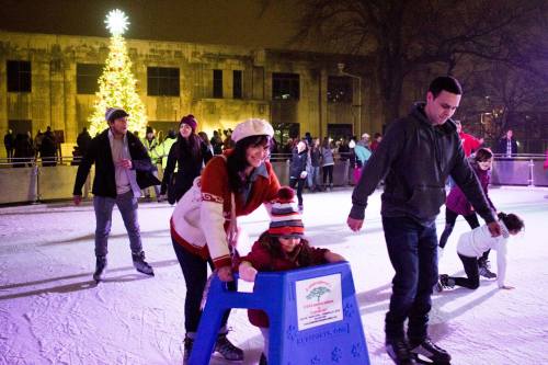 A family enjoys ice skating at night, with a Christmas tree in the background and people skating around them.
