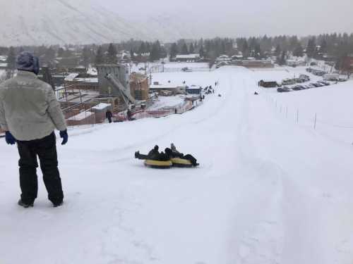 Two people slide down a snowy hill on a tube while another person watches from the side in a winter landscape.