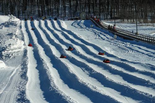 A snowy hill with multiple tubing lanes, people sliding down on inflatable tubes, surrounded by trees and a wooden bridge.
