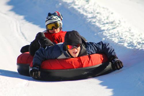 Two children sledding down a snowy hill on a red inflatable tube, wearing winter gear and goggles.
