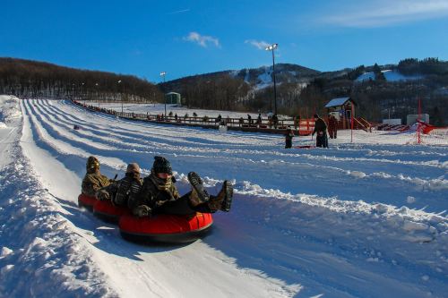 Three people in winter gear slide down a snowy hill on a red tube, with a clear blue sky and trees in the background.