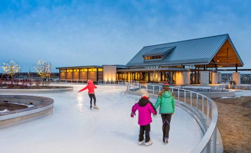 Two children skate on an outdoor rink near a modern building, surrounded by winter lights and a cloudy sky.