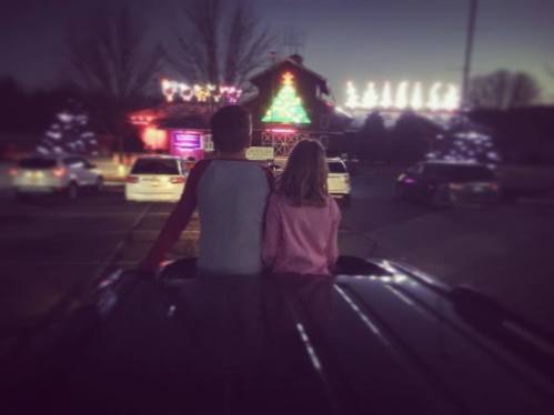 Two children sit on the roof of a car, watching colorful holiday lights in the distance at dusk.