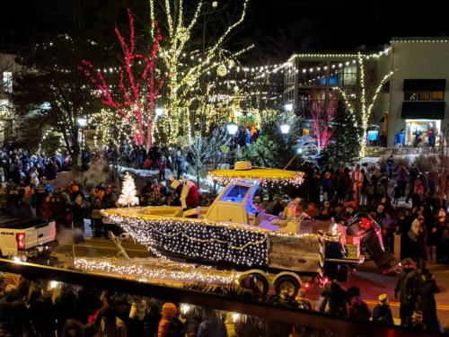 A festive boat float adorned with lights in a crowded parade, surrounded by holiday decorations and cheering spectators.