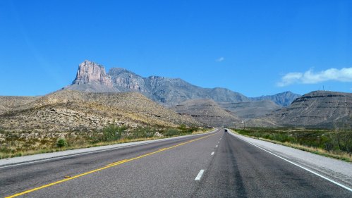 A wide highway stretches toward distant mountains under a clear blue sky, surrounded by arid landscape.