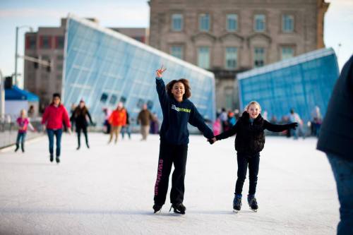 Two children skate joyfully on an ice rink, holding hands, with people and modern buildings in the background.