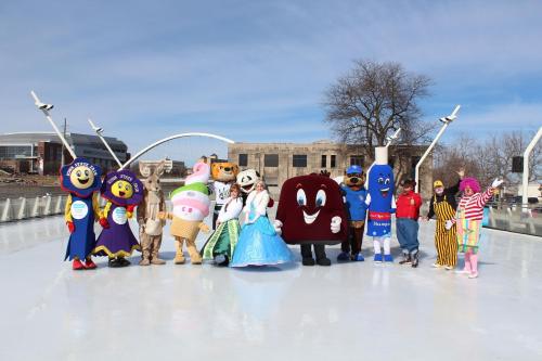 A group of colorful mascots poses on an ice rink, with a clear blue sky and buildings in the background.