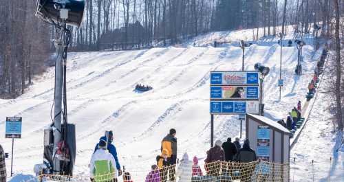 A snowy hill with people tubing down and a ski lift in the background, surrounded by trees and a sign for the tubing area.