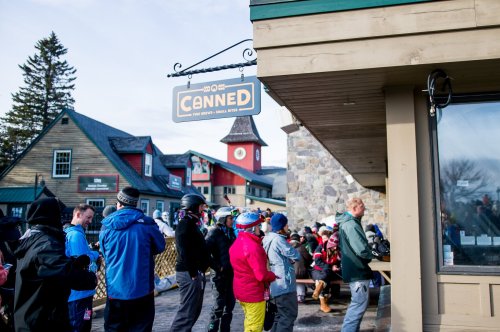 A crowd of skiers and snowboarders gathers outside a building with a sign reading "Canned" in a snowy resort area.