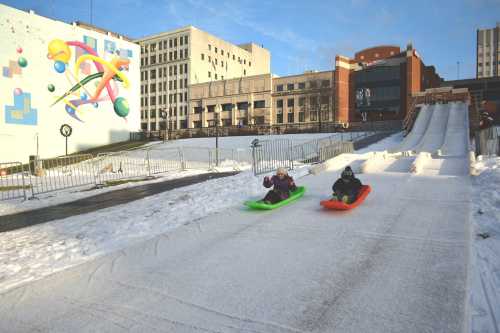 Two children sled down a snowy hill on colorful sleds, with a cityscape and art mural in the background.