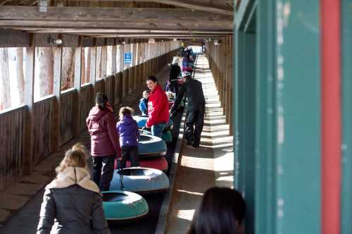 A group of people, including children, waiting in line with colorful tubes for a snow tubing activity in a covered area.