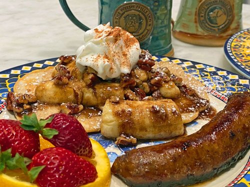 A plate of pancakes topped with bananas, pecans, whipped cream, and a side of strawberries, orange, and sausage.