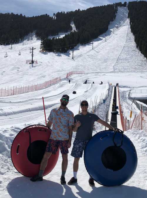 Two men in summer attire stand with snow tubes on a snowy slope, with ski lifts and a mountain in the background.