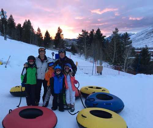 A family of six in winter gear stands on a snowy slope with colorful snow tubes, against a sunset backdrop.