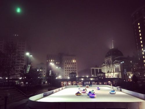 A foggy night scene of a skating rink with colorful bumper cars and city buildings in the background.