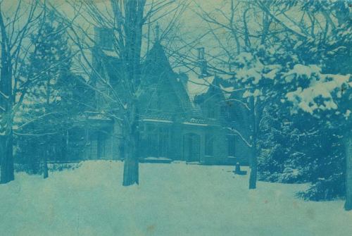 A vintage blue-tinted photo of a snow-covered house surrounded by trees in winter.