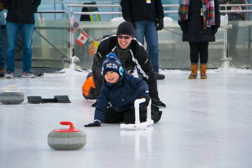 A child in winter gear smiles while sliding on ice, with an adult behind him, both enjoying a curling activity.