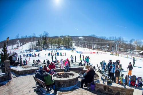 A sunny ski resort scene with people skiing, snowboarding, and relaxing around a fire pit on a snowy slope.