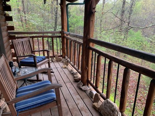 A wooden porch with two chairs and a small table, surrounded by lush greenery and trees.