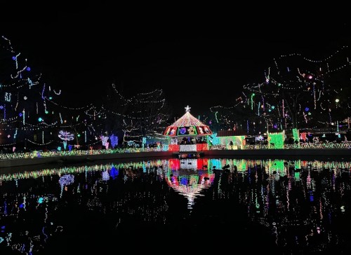 A festive park at night, illuminated with colorful lights reflecting in the water, featuring a decorated gazebo.